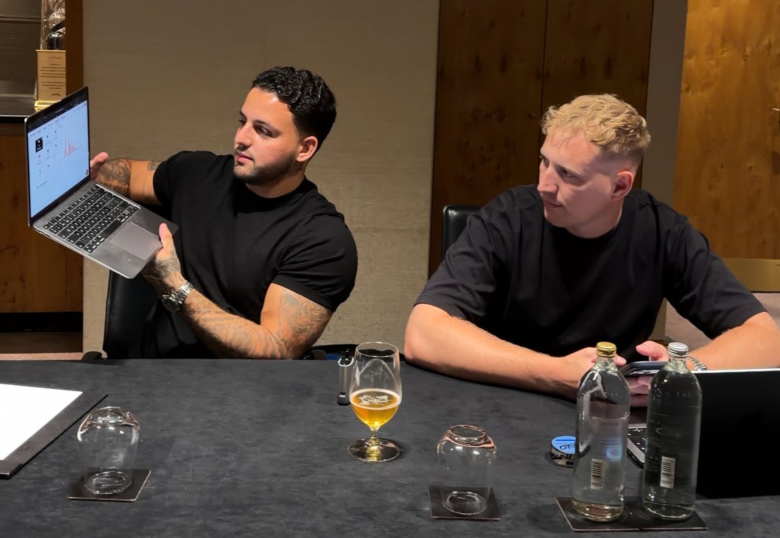 Two men in black t-shirts at a boardroom table, one turning a laptop toward the camera.